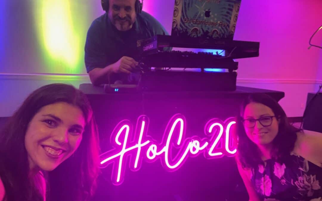 Two people pose in front of a DJ at a table with a neon HoCo20 sign.