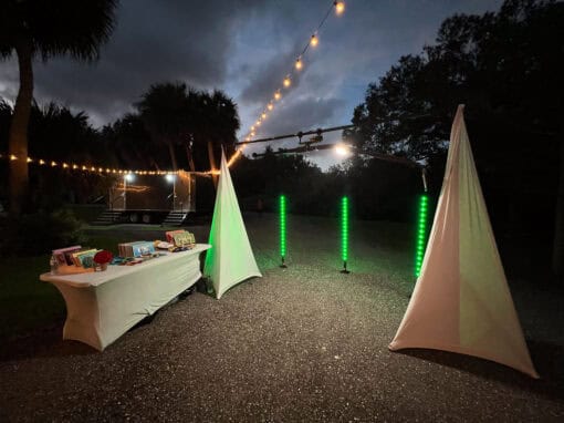 Outdoor nighttime setup with two lit white tents, a table of books, and vertical blue light bars. String lights hang above dark trees and a cloudy sky.