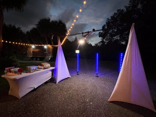 Outdoor nighttime setup with two lit white tents, a table of books, and vertical blue light bars. String lights hang above dark trees and a cloudy sky.