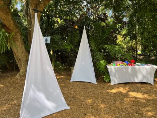 Outdoor area with two white triangular fabric structures and a table with colorful items in the background, surrounded by greenery.