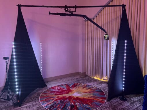 Video booth setup with a circular rug, curtains, two black triangular panels, LED light strips, and a boom arm holding a camera or light.