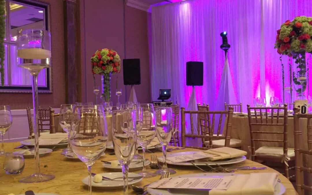 Elegant banquet hall with round tables set with glassware, plates, and floral centerpieces. A purple-lit backdrop and audio equipment are in the background.