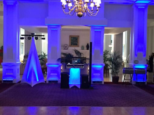 A brightly lit room with blue uplighting features a DJ setup with speakers and equipment beneath a chandelier.