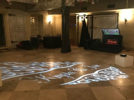 An empty indoor space with a light projection on the floor reading Karyita and Alex. A DJ booth and speakers are set up nearby.