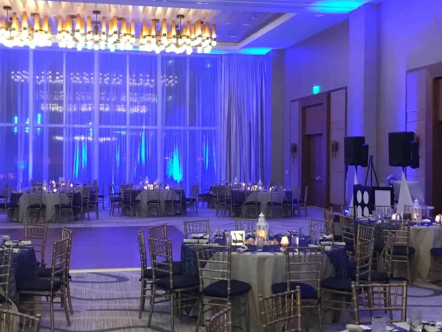 A dimly lit banquet hall with round tables, chairs, and soft blue lighting. Candles and table settings are arranged on the tables.