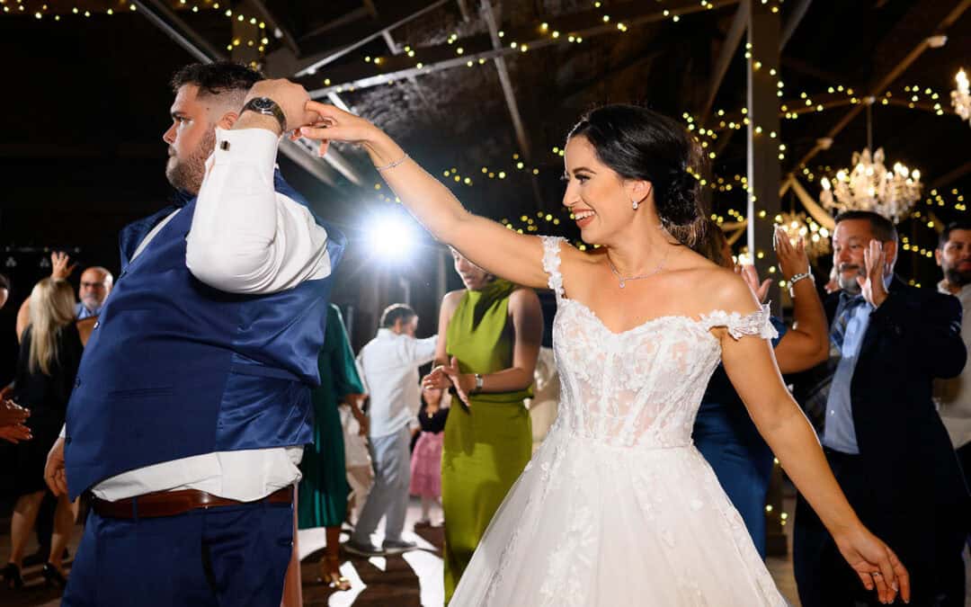 A bride and groom dance joyfully under string lights at a wedding reception, surrounded by guests.