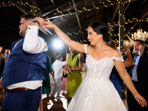 A bride and groom dance joyfully under string lights at a wedding reception, surrounded by guests.