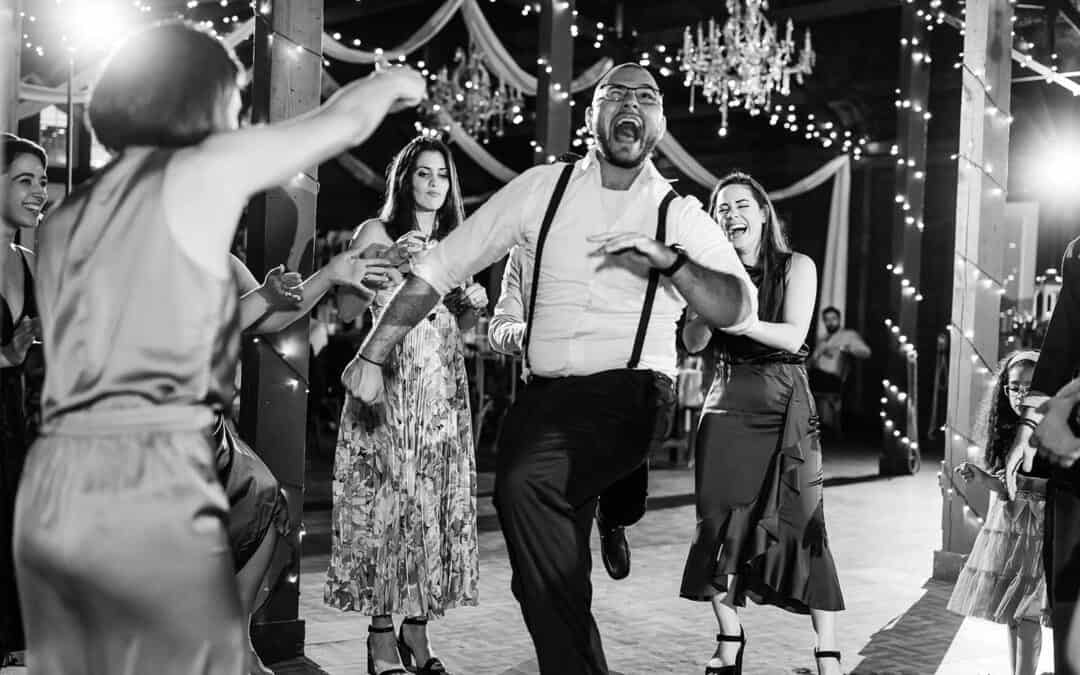 A joyful man in suspenders dances surrounded by smiling people at a festive event, with string lights and chandeliers in the background.