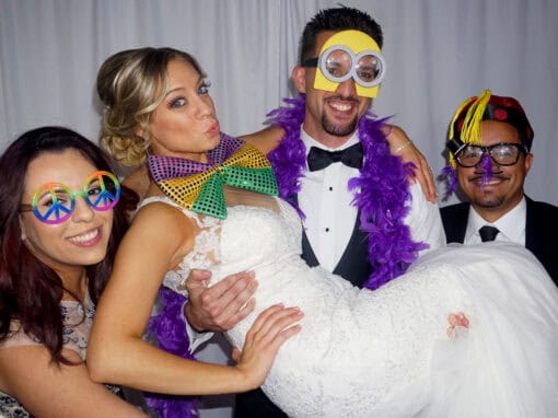 Four people pose playfully; a bride is lifted by a groom and two friends. They wear colorful, quirky accessories over formal attire.