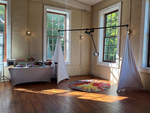 An indoor space with wooden floors, two hanging white drapes, a colorful circular rug, and a table with various items. Sunlight streams through tall windows.