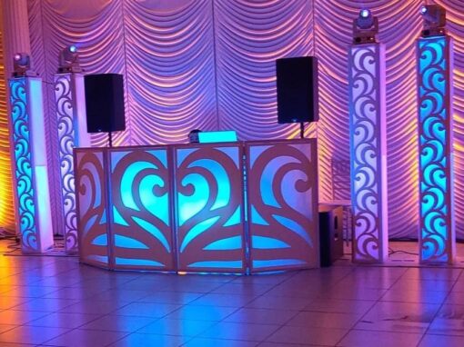 A DJ booth with blue and purple lighting is set against a draped curtain backdrop. Speakers and decorative panels are arranged symmetrically.