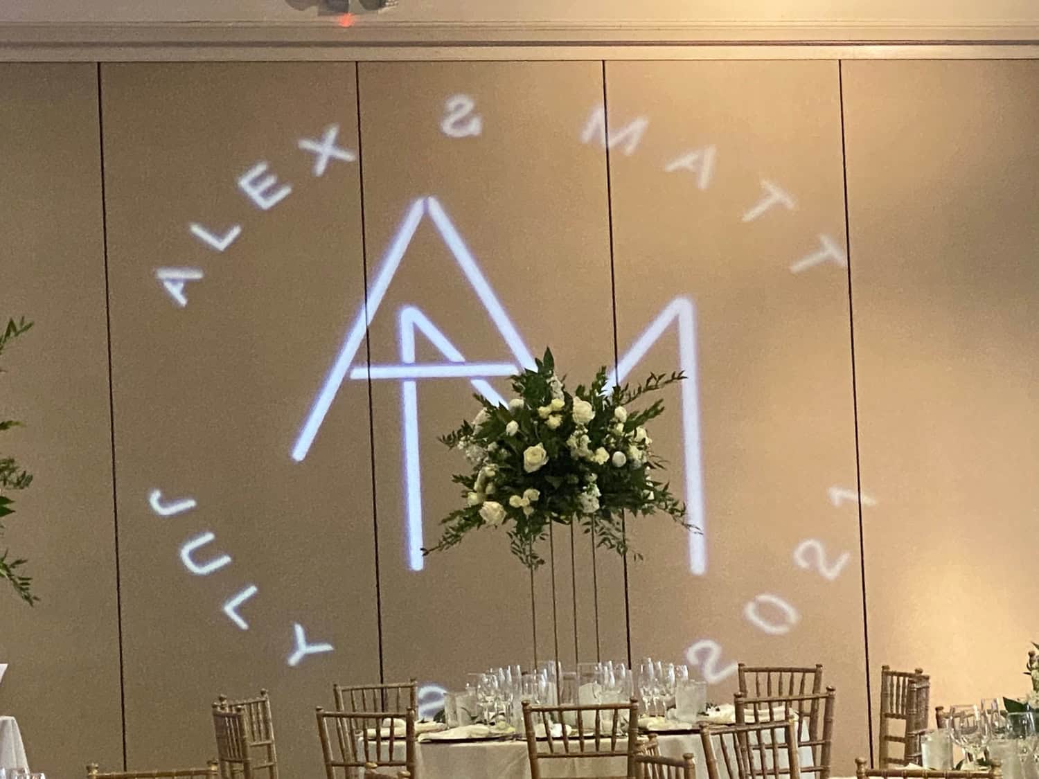 Wedding reception with monogram projection Alex and Matt July 2021 on the wall, with floral centerpiece and table settings.
